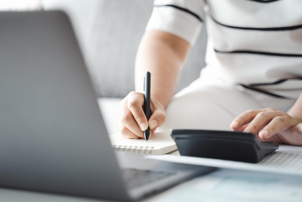 woman wearing white shirt writing on paper in front of a laptop