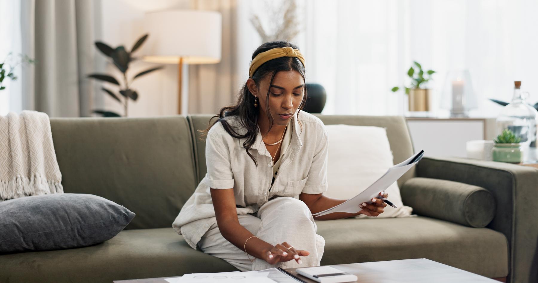 Woman, calculator and budget on sofa of home finance, taxes documents and income or salary management. Young person with paperwork, invoice or loan, inflation and bills for rent cost in living room