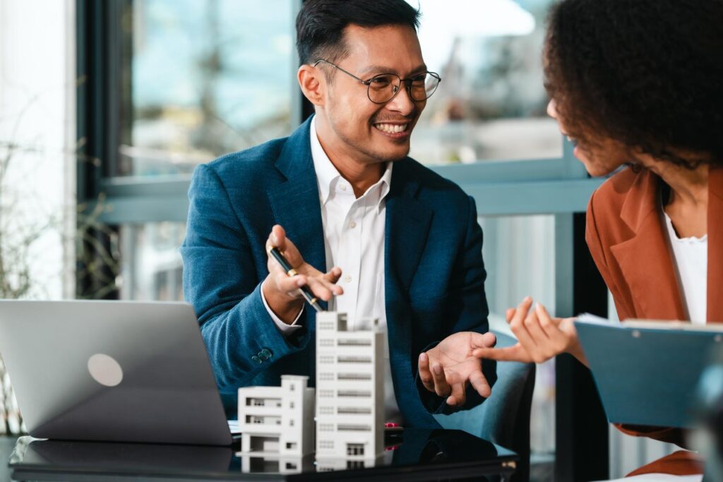 Attractive Asian businessman and African American female real estate and land investor talk discussing together on table with laptop and small building model. Reserve fund.