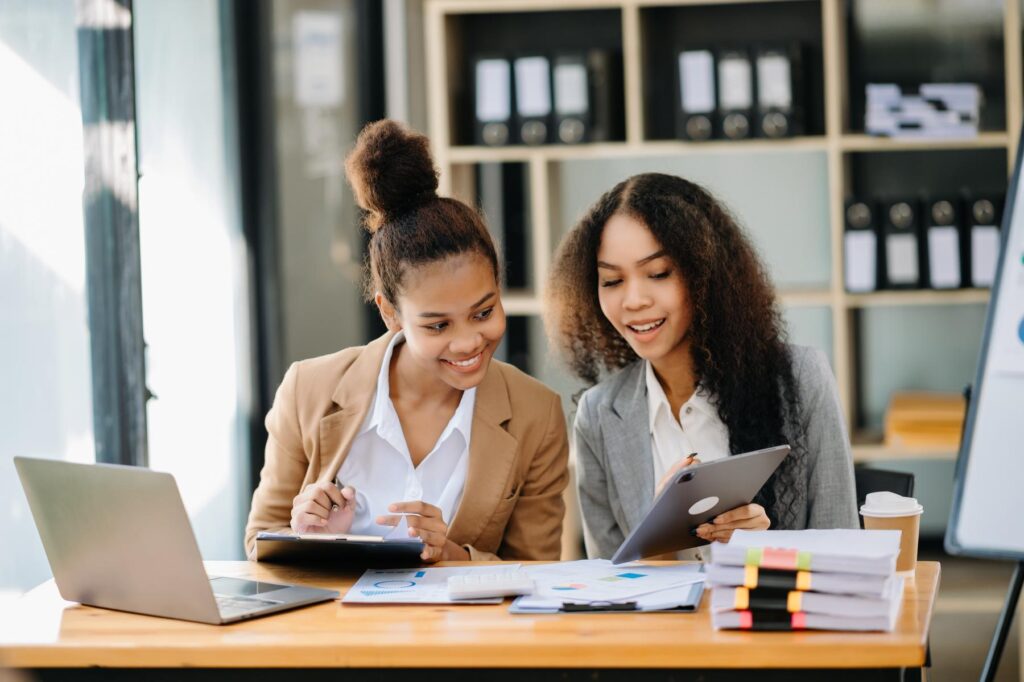 Two African businesswoman discuss investment project working and planning strategy with tablet laptop computer in office.