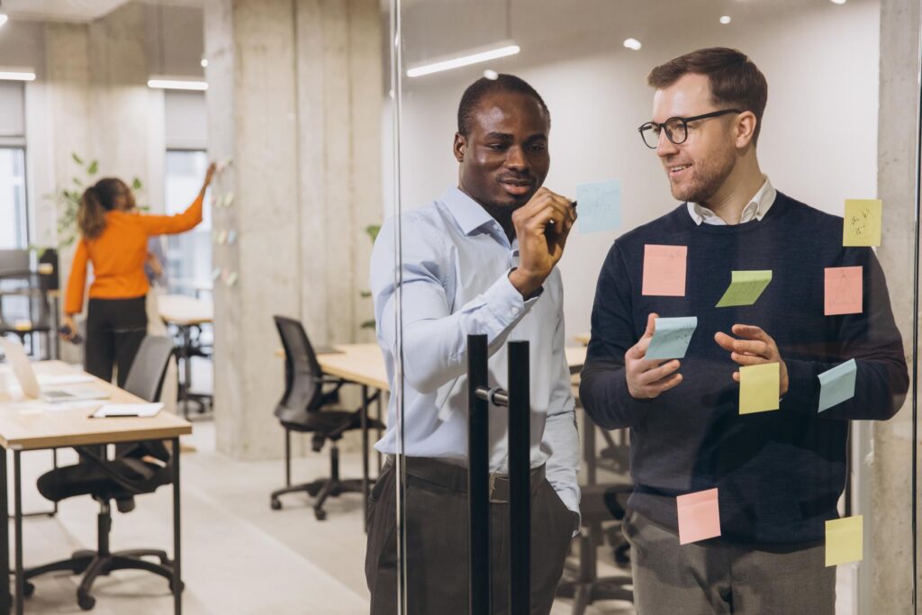 Two businessmen discussing ideas with sticky notes on glass wall, while a colleague works in the background, showcasing teamwork and innovation