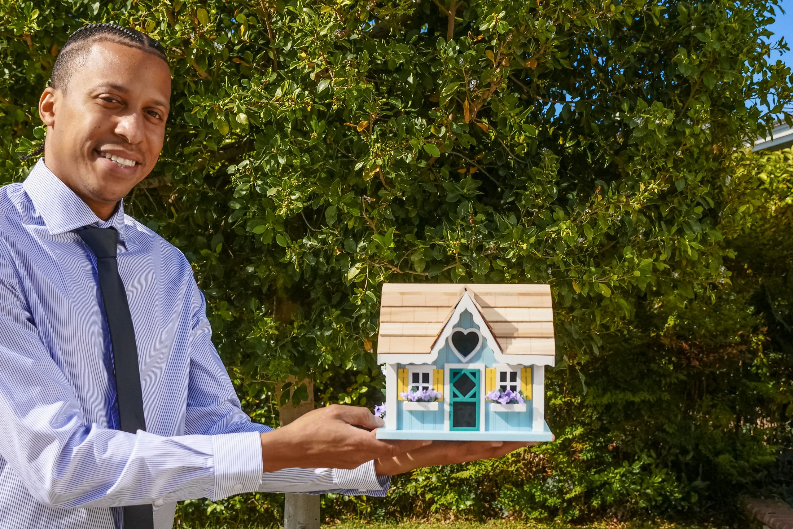 Smiling property manager presenting a miniature house model outdoors against a background of greenery.