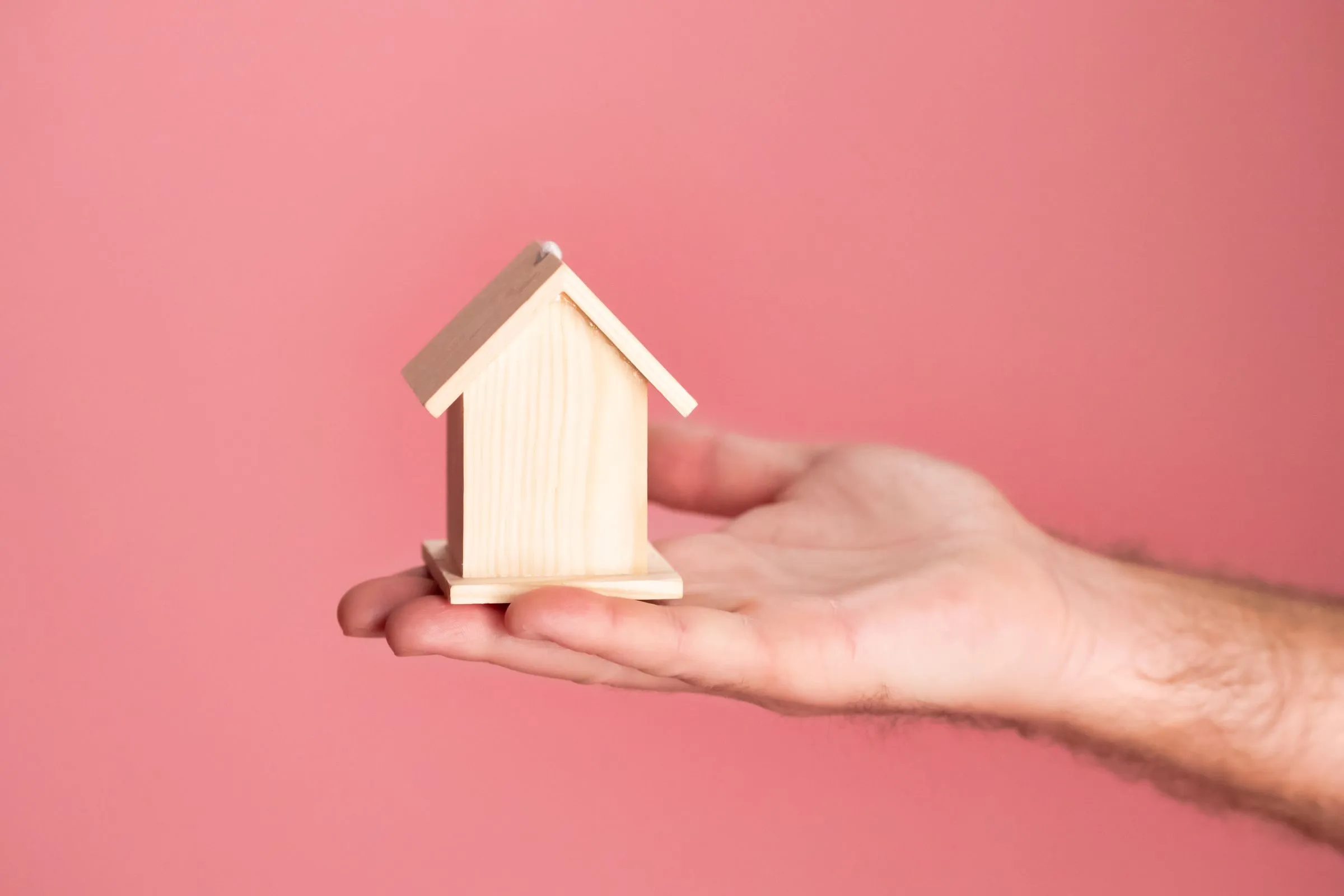 A licensed property manager holding a small wooden house model against a soft pink background, symbolizing real estate and property management.