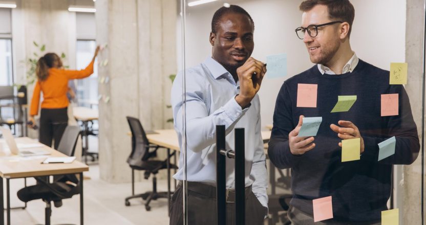 Two businessmen discussing ideas with sticky notes on glass wall, while a colleague works in the background, showcasing teamwork and innovation