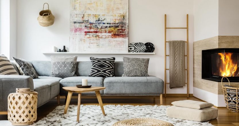 Wooden table next to grey corner settee in warm living room interior with painting and fireplace. Real photo