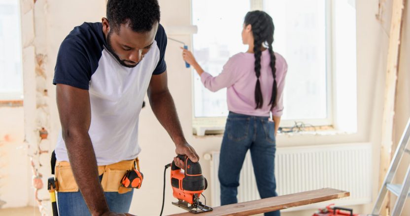 A man cuts a wooden board with a circular saw while a woman paints a wall behind him, handling the rehab stage of the BRRRR method.
