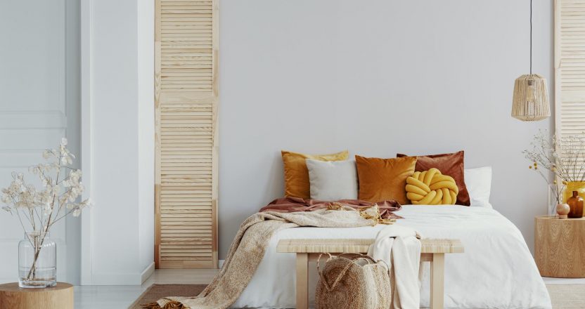 Brown and orange pillows on white bed in natural bedroom interior with wicker lamp and wooden bedside table with vase