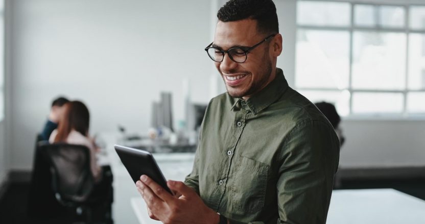 A smiling man in an office looks at a tablet while researching whether real estate is a good investment.