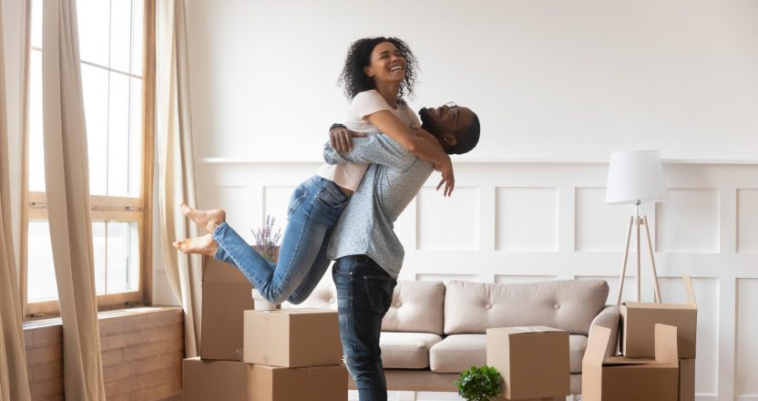 Man lifts a laughing woman in a living room full of moving boxes, illustrating the human side of real estate investing.