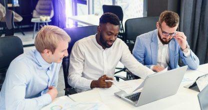 Three businessmen sitting at the modern office tables and analysing data from report and computer to succeed in developing of their own company.