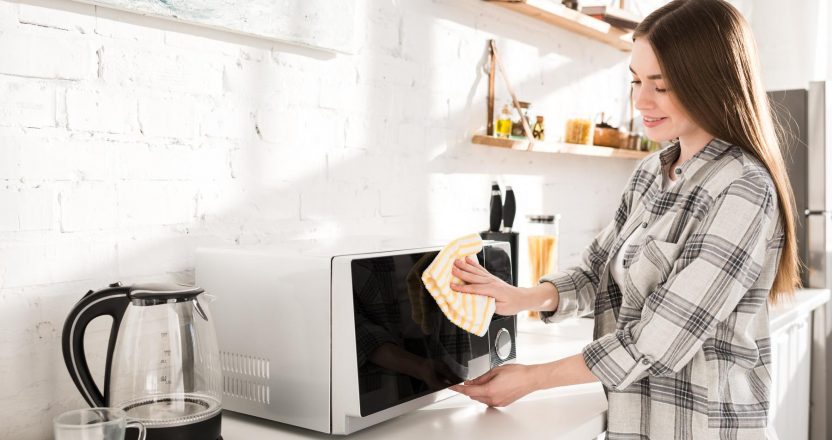 smiling and attractive woman cleaning microwave with rag in kitc