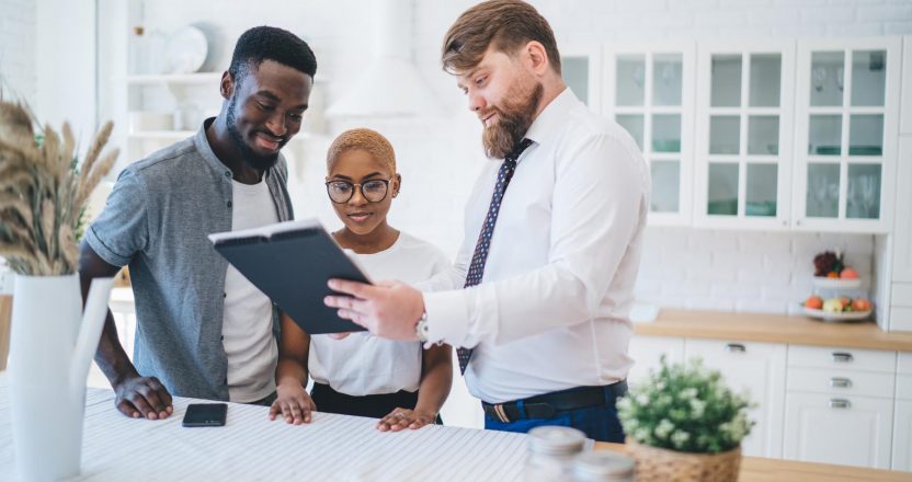 Confident estate agent showing documents to African American happy couple while standing in kitchen