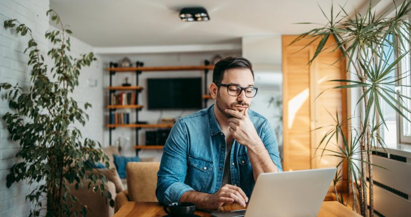 Portrait of a casual businessman thinking while looking at laptop screen, keeping hand on chin.