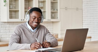 Black male student wearing headphones elearning on laptop at home office.