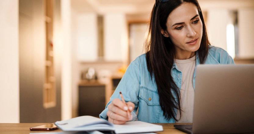 Focused charming woman writing down notes while working with laptop
