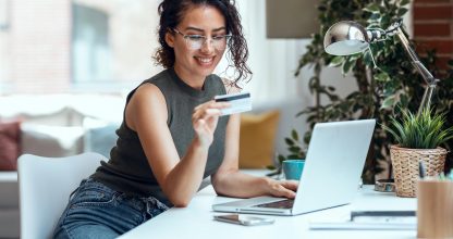 Cute woman holding white credit card for shopping online with computer while sitting in living room at home.
