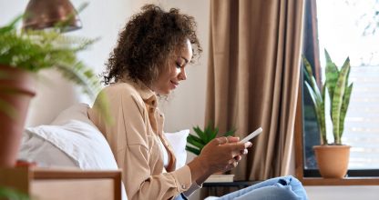 Young happy African American woman using cellphone sitting on bed.