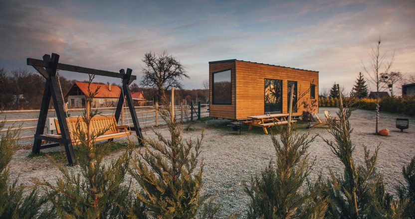 A modern wood-clad tiny home sits on a gravel lot beside a wooden swing set at dusk, one option shaping the cost of tiny homes.