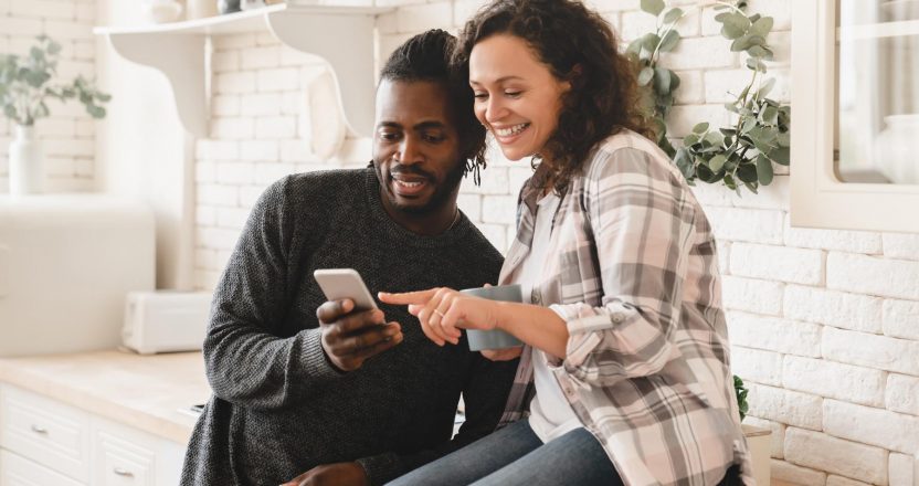 Middle-aged mature happy african couple using smart phone cellphone for surfing social media, sharing photos, checking e-mail, using mobile application online relaxing at home kitchen