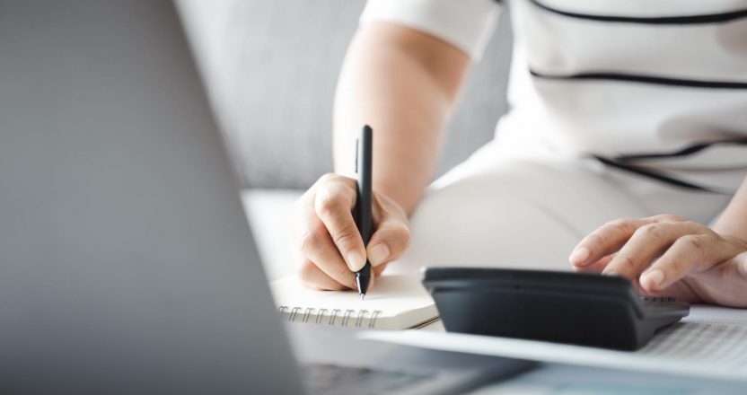woman wearing white shirt writing on paper in front of a laptop