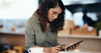 Small business owner or entrepreneur filling out paper work or a document in an internet cafe or coffee shop with a phone in hand. Young woman writing on a from and sitting at a table in a restaurant