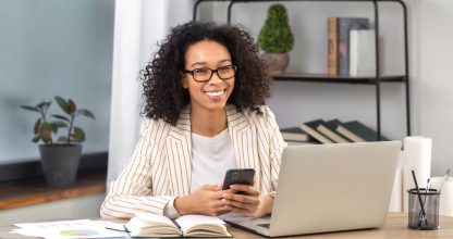 Portrait of young successful African American businesswoman or office worker sitting at a desk with laptop in modern office, looking at the camera and smiling