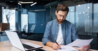 Serious and focused financier accountant on paper work inside office, mature man using calculator and laptop for calculating reports and summarizing accounts, businessman at work in casual clothes