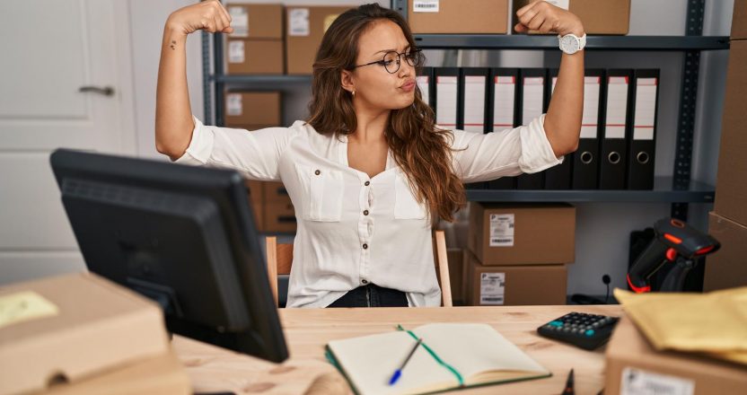 Young hispanic woman working at small business ecommerce showing arms muscles smiling proud. fitness concept.