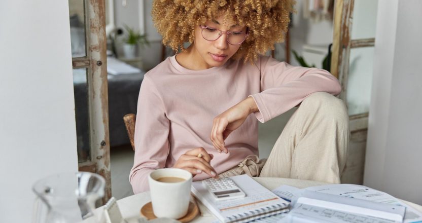 Serious curly haired woman manages household family budget calculates expenditures takes care of finances and savings sits at table with receipts dressed in domestic clothes poses at home office