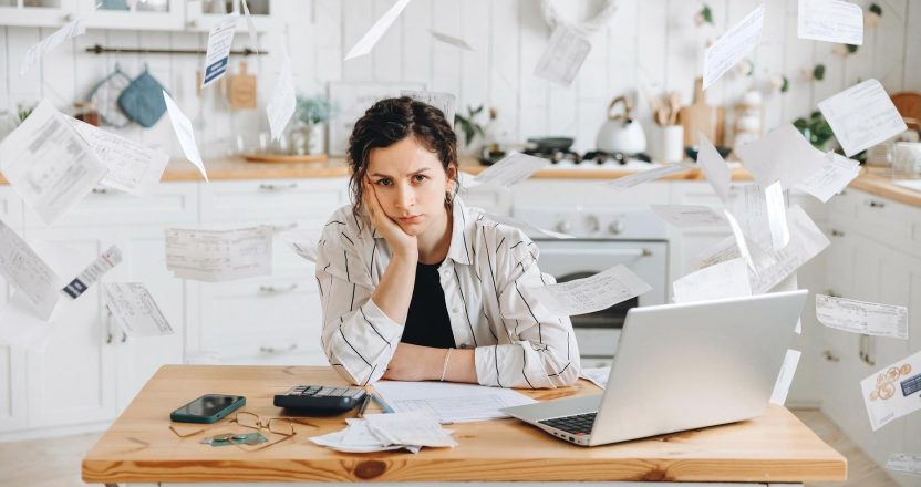 A woman sits overwhelmed as paper bills fly around her kitchen table, tracking receipts from paying rent with Venmo.