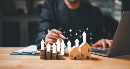 A businessman holding a coin in his hand ponders the concept of wealth and financial security as he looks at a house model, representing the potential investment of a mortgage loan.