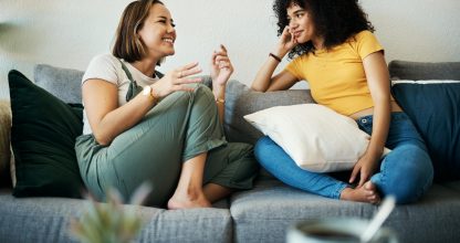 Women, friends relax and conversation in a home with gossip, discussion and happy in a living room. Couch, smile and female person on sofa with communication together in a house lounge with speaking