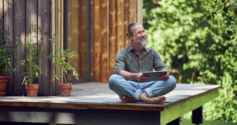 Man with a tablet relaxes barefoot on the porch of a compact wooden home, the kind of build behind most tiny house costs.