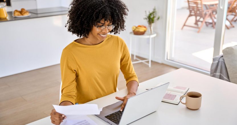 Young smiling happy African woman checking paper paying bills online at home.