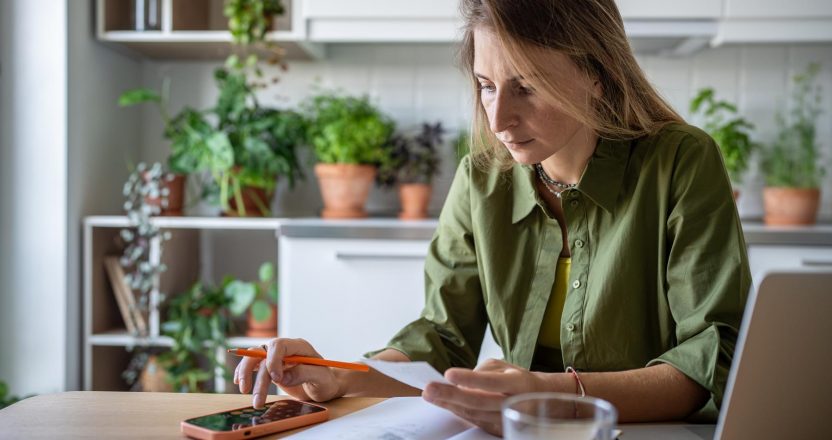 Concentrated female holding bill for utilities payment, checking figures on mobile phone calculator