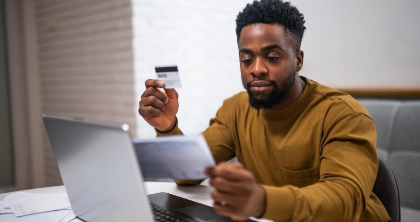 Modern young black man using credit card and laptop at home. He is focusing on e-commerce and managing his finances.