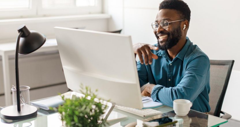 Handsome black male entrepreneur sitting at desk in office, looking at computer monitor and smiling, having video call