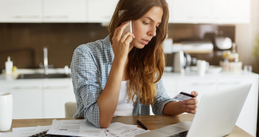 Beautiful young Caucasian woman calling bank using cell phone concerning information on credit card that she is holding. Serious female connecting to mobile banking service using electronic device