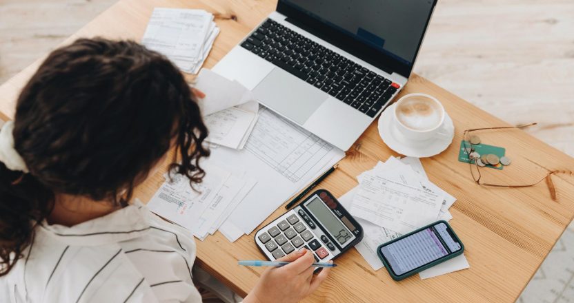 Top view of a woman working at home in the kitchen with financial papers, counting on a calculator, paying bills, planning a budget to save some money. Independent accounting, remote accountant