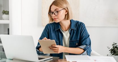 Young woman taking notes using laptop for remote studies at home desk. Smart freelancer watching learning educational webinar, counting charts and finances
