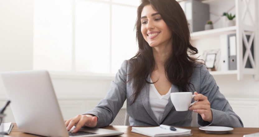 Smiling businesswoman working on laptop and drinking coffee at office