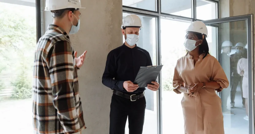 Licensed property managers discuss project indoors wearing masks and hard hats, ensuring safety.