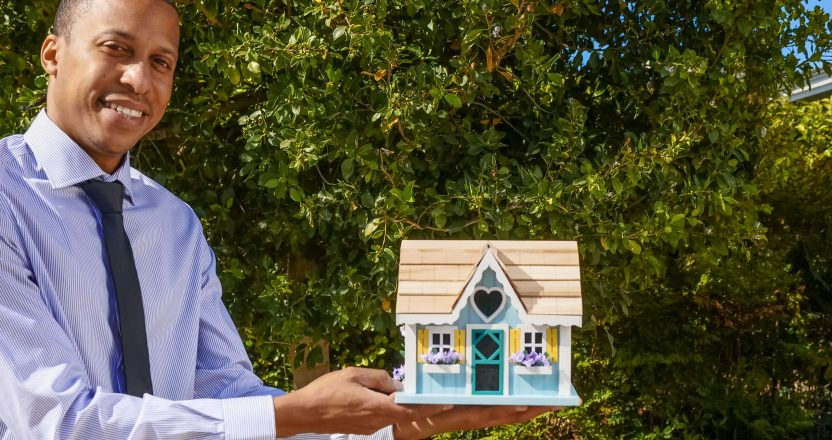 Smiling property manager presenting a miniature house model outdoors against a background of greenery.
